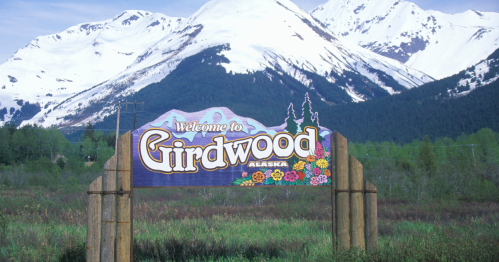 A colorful welcome sign for Girdwood, Alaska, with snow-capped mountains in the background and lush greenery in the foreground.