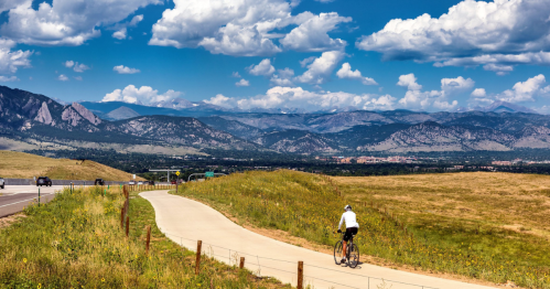 A cyclist rides along a paved path with mountains and blue skies in the background, surrounded by green fields.
