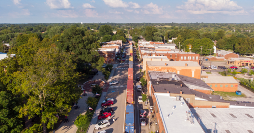 Aerial view of a small town street lined with trees, buildings, and parked cars under a partly cloudy sky.