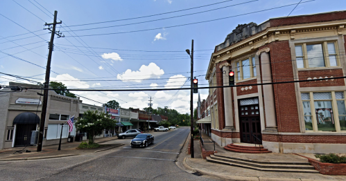A street view of a small town with a brick building, traffic lights, and shops lining the road under a cloudy sky.