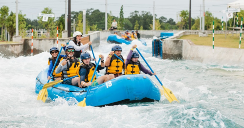 A group of people in yellow life jackets paddling a blue raft through whitewater rapids in a water sports facility.