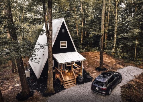 A black A-frame cabin nestled in a wooded area, with a gravel driveway and a parked car nearby.