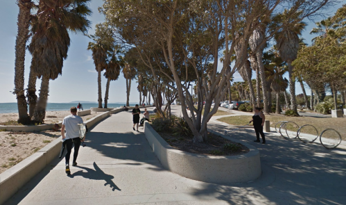 A sunny beachside walkway lined with palm trees, with people walking and bicycles parked nearby.