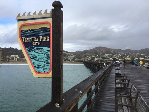 Sign for Ventura Pier, established in 1872, with a wooden pier extending over the water and hills in the background.