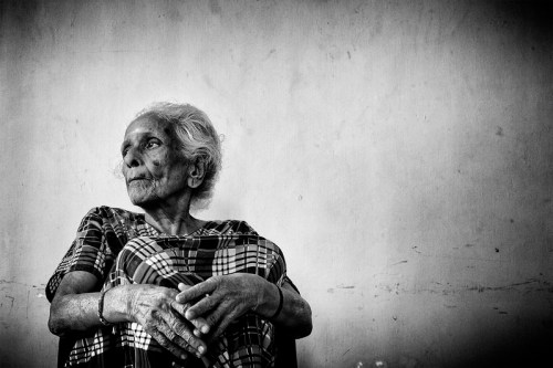 An elderly woman in a patterned sari sits thoughtfully against a plain wall, captured in black and white.