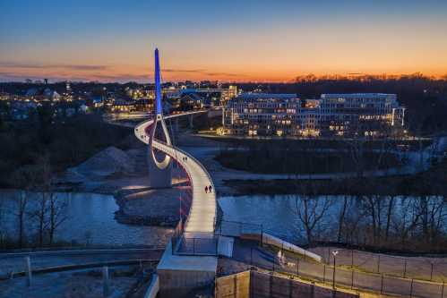 A modern bridge with a spiral design spans a river, illuminated at dusk, with buildings and a sunset in the background.