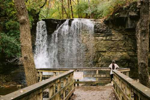 A person stands on a wooden bridge, gazing at a waterfall surrounded by lush greenery and rocky cliffs.