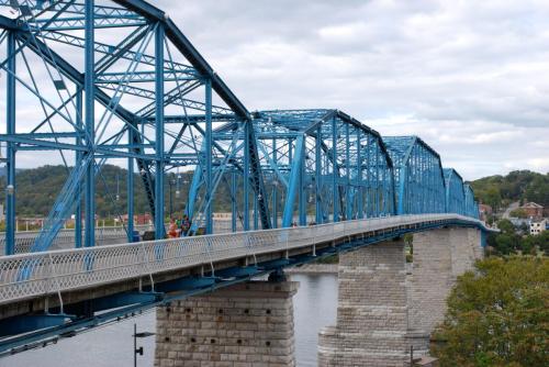 A blue steel bridge spans a river, with stone supports and a cloudy sky in the background.