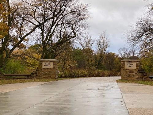Entrance to Lake Arounabi State Park, featuring stone pillars and a paved path surrounded by autumn foliage.