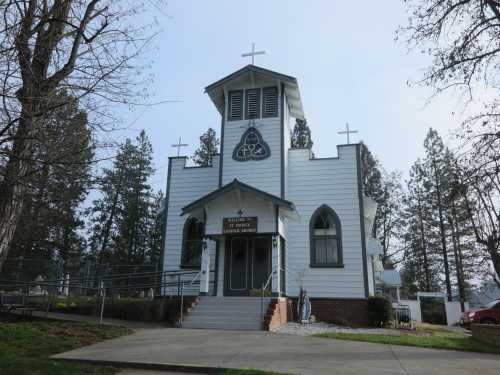 A white church with a triangular roof and multiple crosses, surrounded by trees and a clear sky.