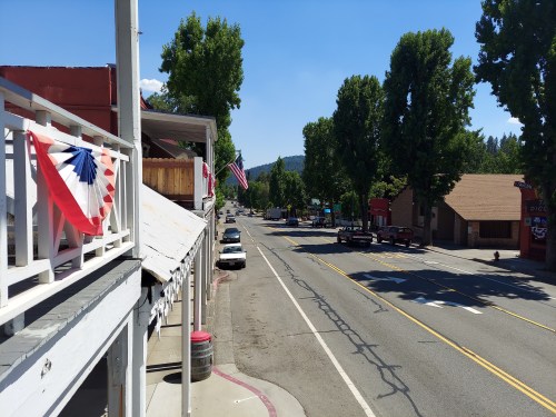 A quiet street scene with trees, parked cars, and patriotic decorations on buildings under a clear blue sky.