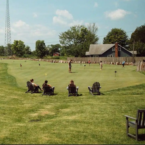 A sunny golf course scene with people putting, surrounded by trees and a house in the background, with chairs in the foreground.