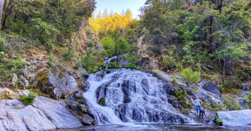 A serene waterfall cascades over rocks, surrounded by lush greenery and trees under a clear blue sky.