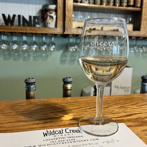 A glass of white wine on a bar counter with a "cheers" inscription, alongside a menu for Wildcat Creek Winery.
