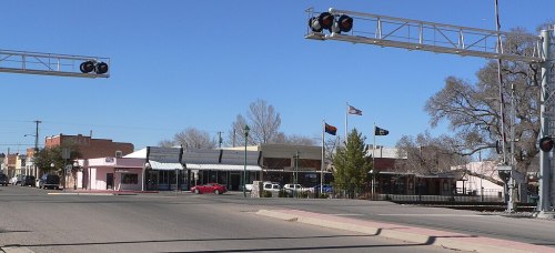 A street view featuring a railroad crossing, shops, and flags in a small town under a clear blue sky.