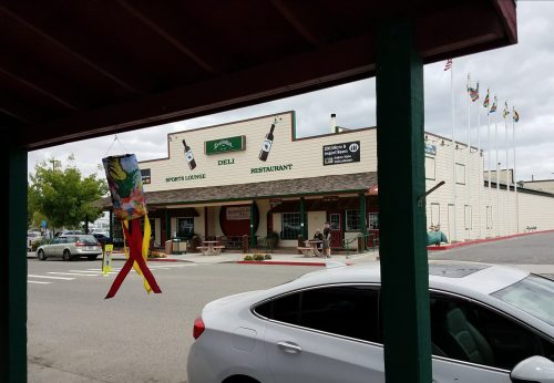 A view of a deli and sports lounge with flags, outdoor seating, and a parked car in front.
