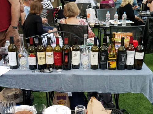A table with various wine bottles lined up, set in a casual outdoor gathering with people in the background.