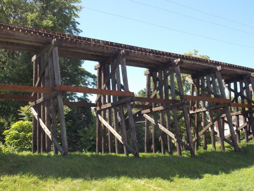 A wooden railway trestle bridge supported by tall wooden beams, surrounded by greenery under a clear blue sky.