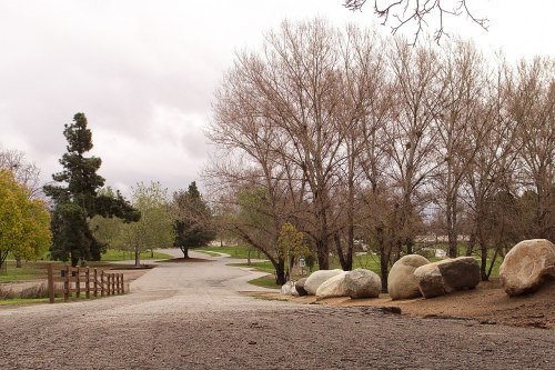 A gravel path leads through a park with bare trees and large rocks, under a cloudy sky.