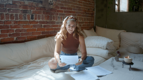 A young woman sits on a bed, holding papers, with a warm smile and sunlight streaming through a window.