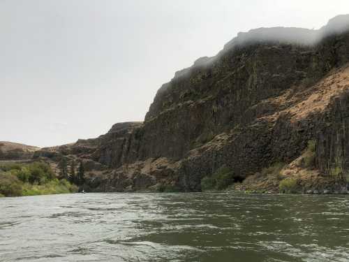 A river flows past steep, rocky cliffs under a cloudy sky, with greenery along the water's edge.