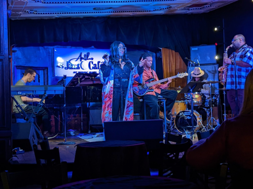 A live music performance at a café featuring a singer, keyboardist, bassist, and saxophonist on stage.