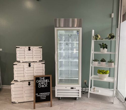A display area featuring a glass refrigerator, stacked crates, and a sign for "The Caramel Apple Chick" in a green room.