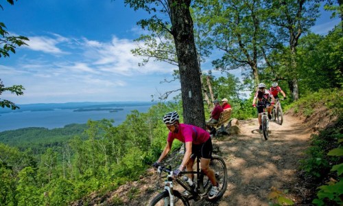 Mountain bikers ride along a scenic trail with a lake view, surrounded by lush trees and a clear blue sky.