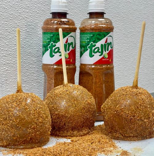 Three caramel apples coated in Tajín seasoning, with two bottles of Tajín in the background.