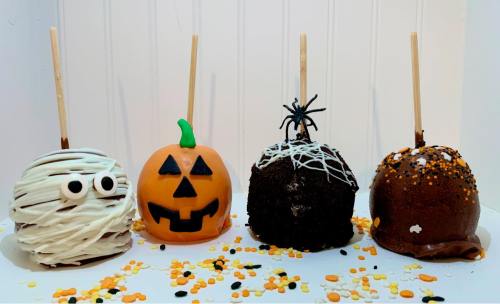 Four decorated Halloween caramel apples: a mummy, a pumpkin, a spider, and a ghost, surrounded by colorful sprinkles.
