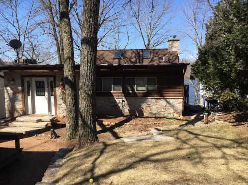 A cozy cabin by a lake, featuring a stone exterior, large windows, and trees casting shadows on the lawn.