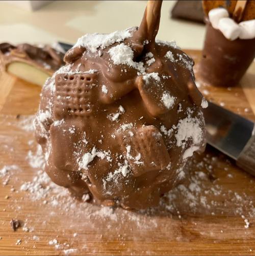 A chocolate-covered dessert topped with powdered sugar and cookie pieces, resting on a wooden board.