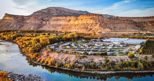 Aerial view of a campground by a river, surrounded by mountains and greenery, with RVs parked in a circular layout.