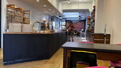 A cozy café interior with a counter, baked goods display, and a customer in a colorful shirt. Tables are set for patrons.