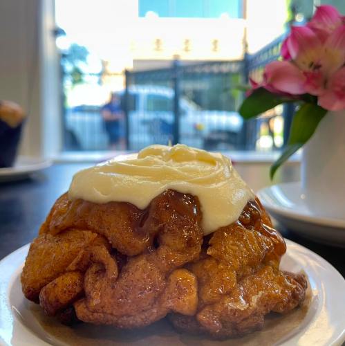 A close-up of a cinnamon roll topped with cream cheese frosting, sitting on a plate with a flower in the background.