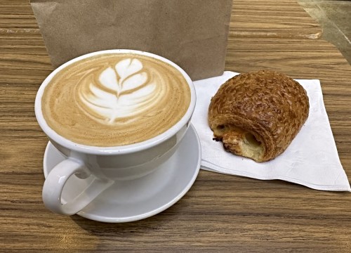 A cup of latte with a leaf design, next to a croissant on a napkin, on a wooden table.