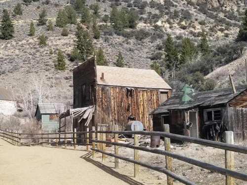 Abandoned wooden buildings in a rural landscape, surrounded by hills and sparse trees. A dirt path leads in front.
