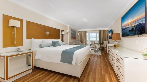 A modern hotel room featuring a king bed, TV, dresser, and seating area with large windows and warm wood flooring.