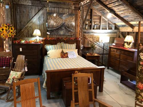 Cozy rustic bedroom display with wooden furniture, warm lighting, and autumn decorations in a barn setting.