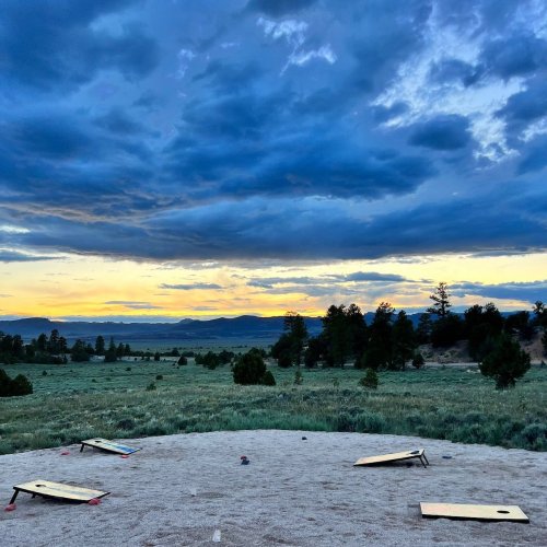 A scenic view of a grassy field with cornhole boards set up, under a dramatic sky at sunset.