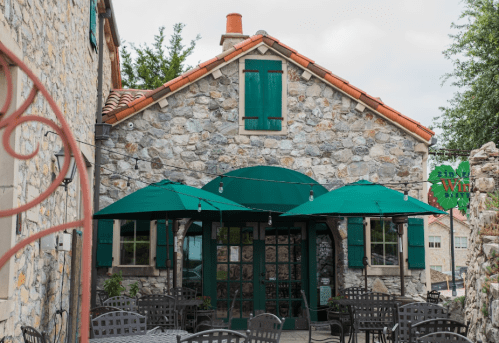 A charming stone building with green shutters and awnings, featuring outdoor seating under umbrellas.