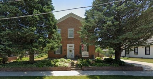 A brick house with large trees in front, featuring a sign and an American flag, set in a green landscape.