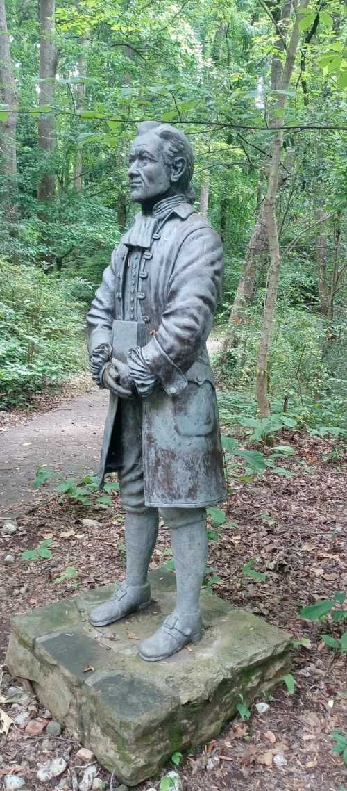 A bronze statue of a man in period clothing stands on a stone base, surrounded by trees and a forest path.