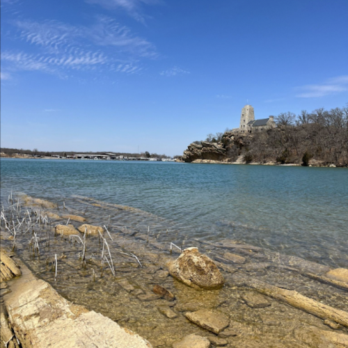 A serene lake scene with clear water, rocky shore, and a distant building on a hill under a blue sky.