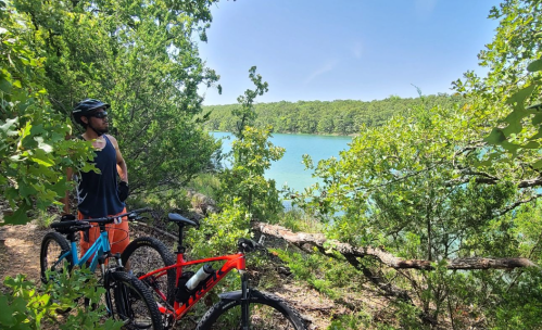 A person stands near a lake, surrounded by trees, with two mountain bikes resting beside them.