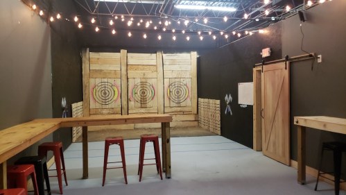 An indoor axe-throwing venue with wooden targets, stools, and string lights overhead.