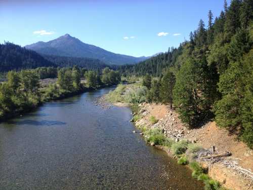 A serene river flows through a lush green landscape, surrounded by mountains under a clear blue sky.