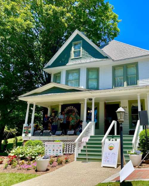 A charming white and green house with a porch, surrounded by trees and flowers, hosting a small outdoor event.