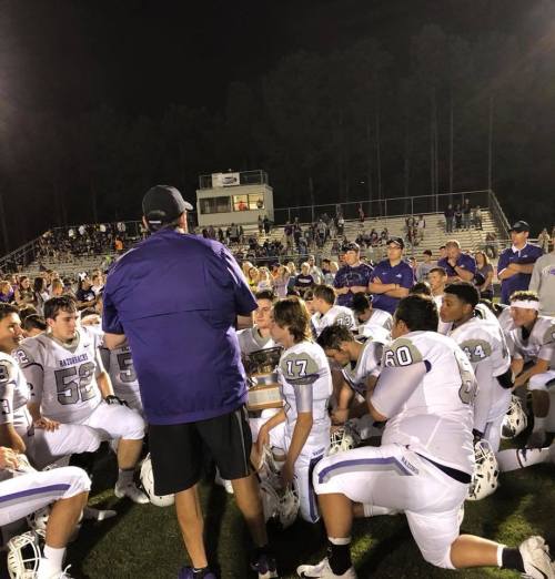 A football coach addresses his team after a game, with players seated around him and a trophy in his hands.