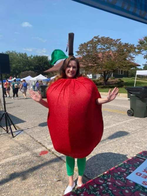 A person in a large red apple costume stands with arms raised at an outdoor event, smiling in a festive atmosphere.